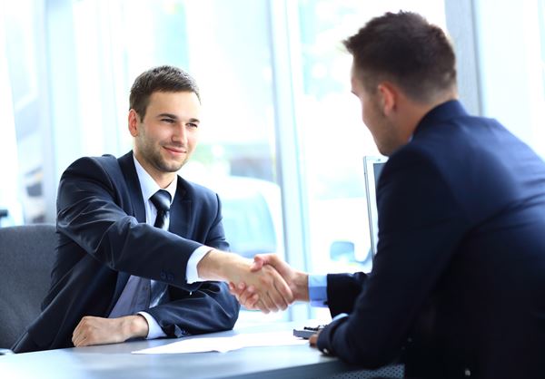 Two people in blue suits smiling and shaking hands across a desk in an office: recruitment industry