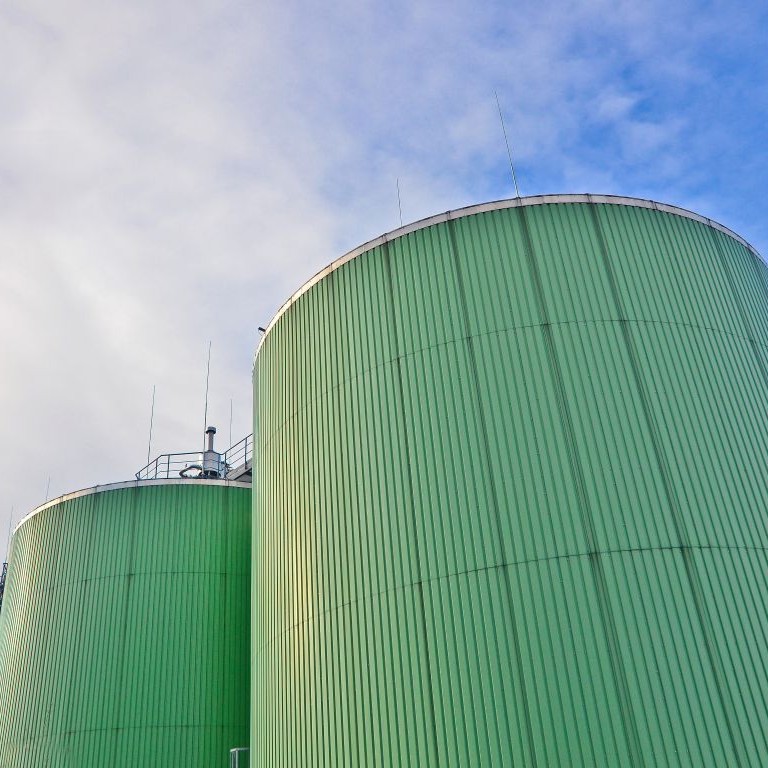 Anaerobic digestion plant with three large green cylindrical digesters, external stairway and cloudy sky