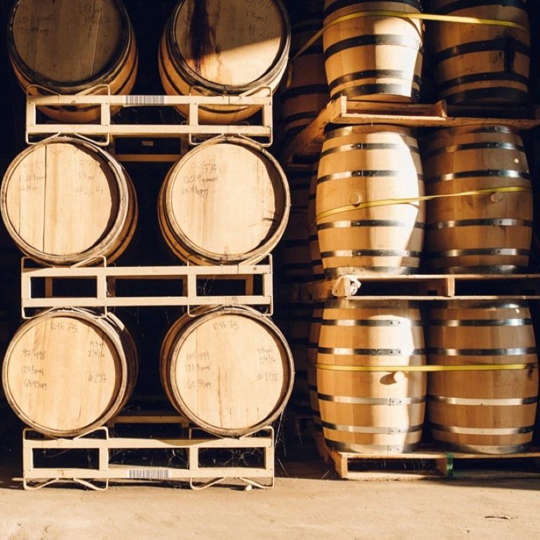 Interior of whiskey maturation warehouses with rows of stacked oak barrels on metal and wooden racks; warm sunlight highlights the barrel staves and handwritten batch markings for aging.