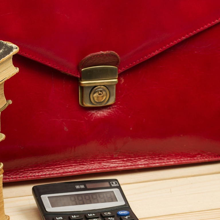 Red leather briefcase with a lock, stacked old books, and a calculator on a wooden surface.