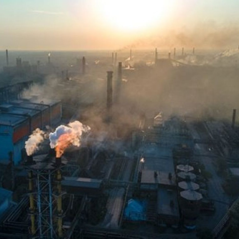 Construction site with industrial buildings, chimneys, and visible smoke or steam in a hazy sunrise scene.