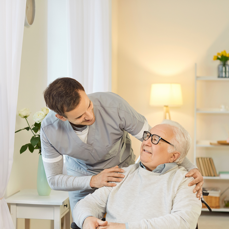 A caregiver in a light blue uniform gently supporting an elderly person seated in a wheelchair in a bright, home-like room with soft natural light and plants.