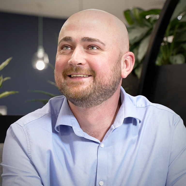 A bald man smiling, wearing a light blue dress shirt is seated indoors with plants and a hanging light bulb in the background.