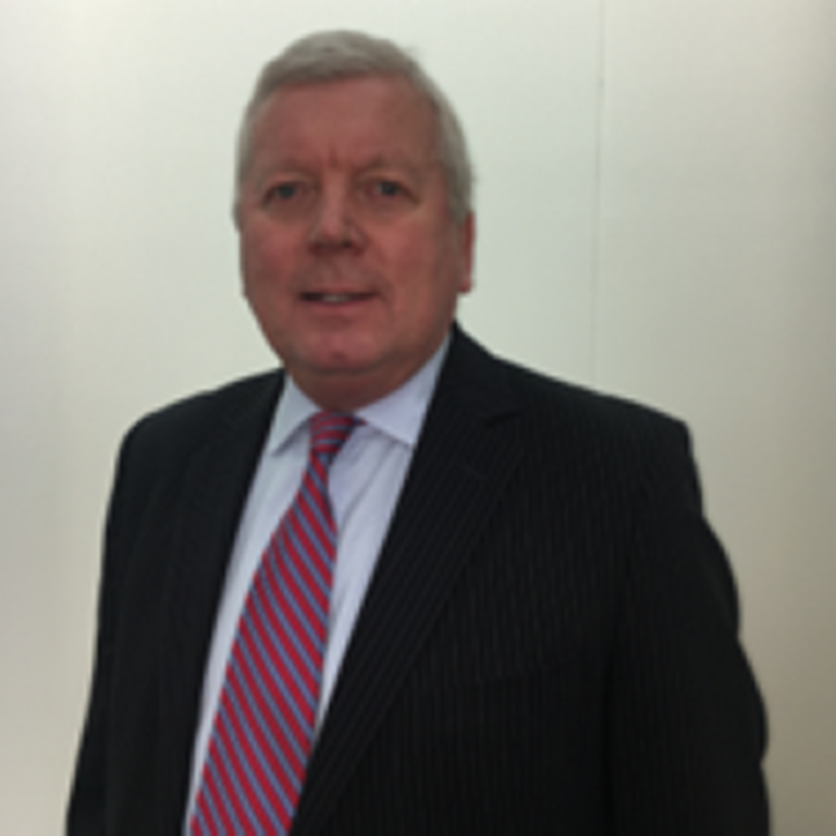 Man wearing a dark suit jacket, white shirt, and red-striped tie standing against a plain light-colored wall.