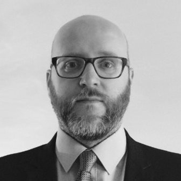 Man wearing a dark suit, light-colored shirt, and patterned tie against a plain background.