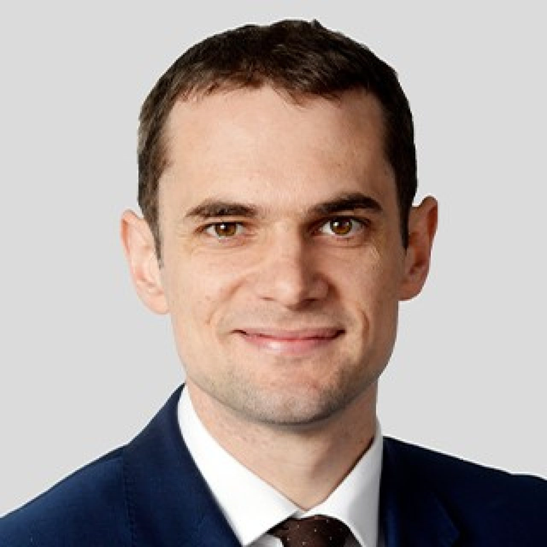 James Crask wearing a dark suit and tie, standing against a light grey background, presenting a professional and approachable appearance.