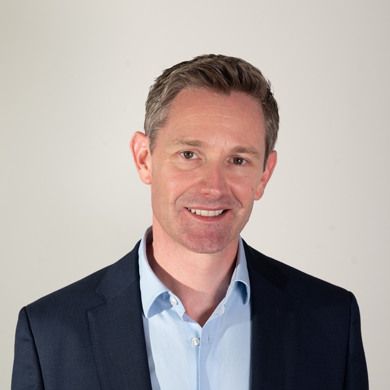 A man in a navy blazer and light blue shirt, posing against a plain light background.