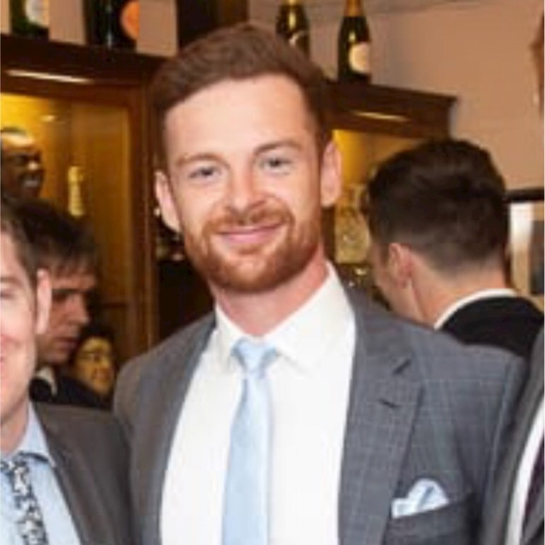 Jamie Roskell dressed in a grey checked suit with a white shirt and light blue tie, standing in a social setting with other people in the background.