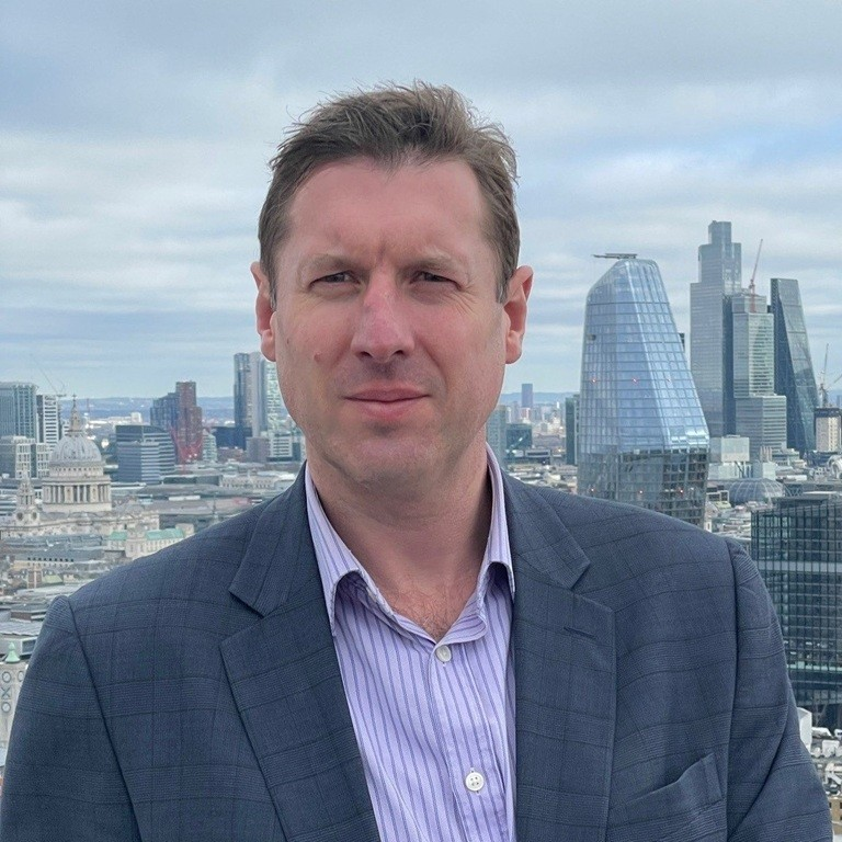 A man wearing a blue checkered blazer and a light purple striped shirt standing outdoors with the London skyline, including modern skyscrapers and historic buildings, visible behind him.