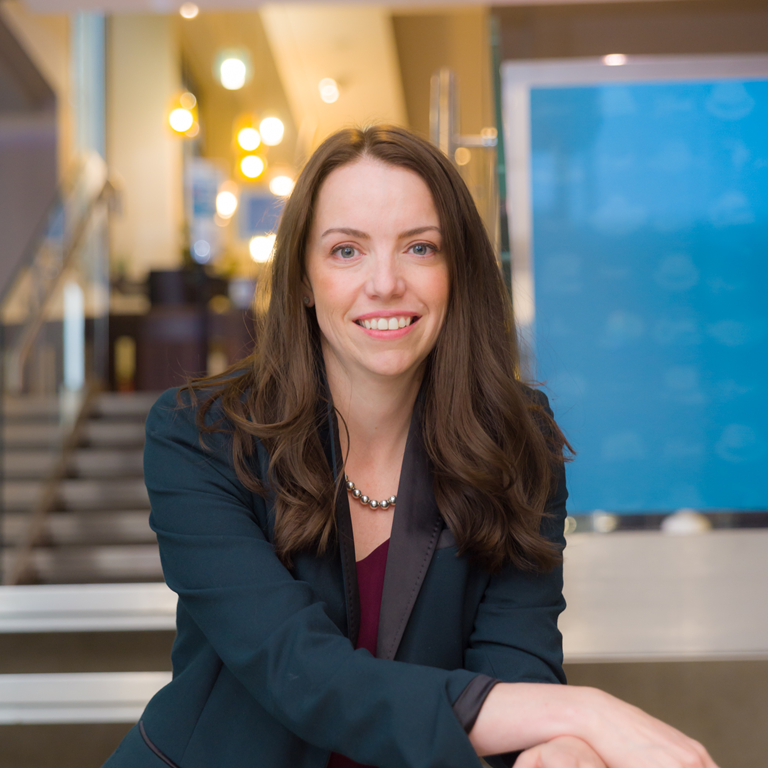 A woman with long brown hair wearing a dark blazer and a pearl necklace, adjusting her sleeve while seated indoors with a staircase and warm lighting in the background.