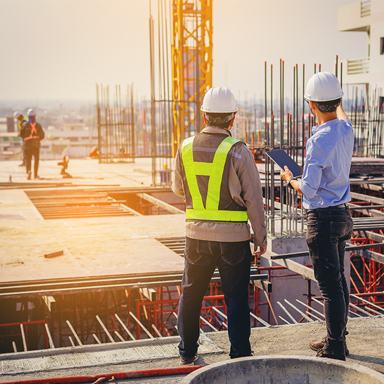 Two construction workers wearing safety helmets and vests discussing plans on a building site with steel reinforcements and scaffolding in the background, illustrating trade credit in construction projects