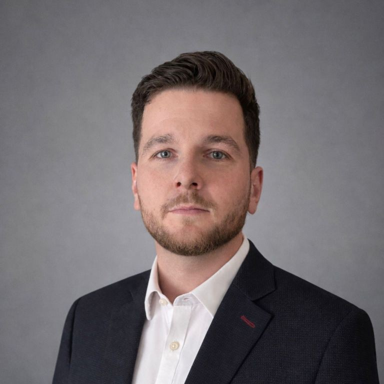 Portrait of Miles Rosewarne wearing a dark blazer and white shirt against a plain gray background.