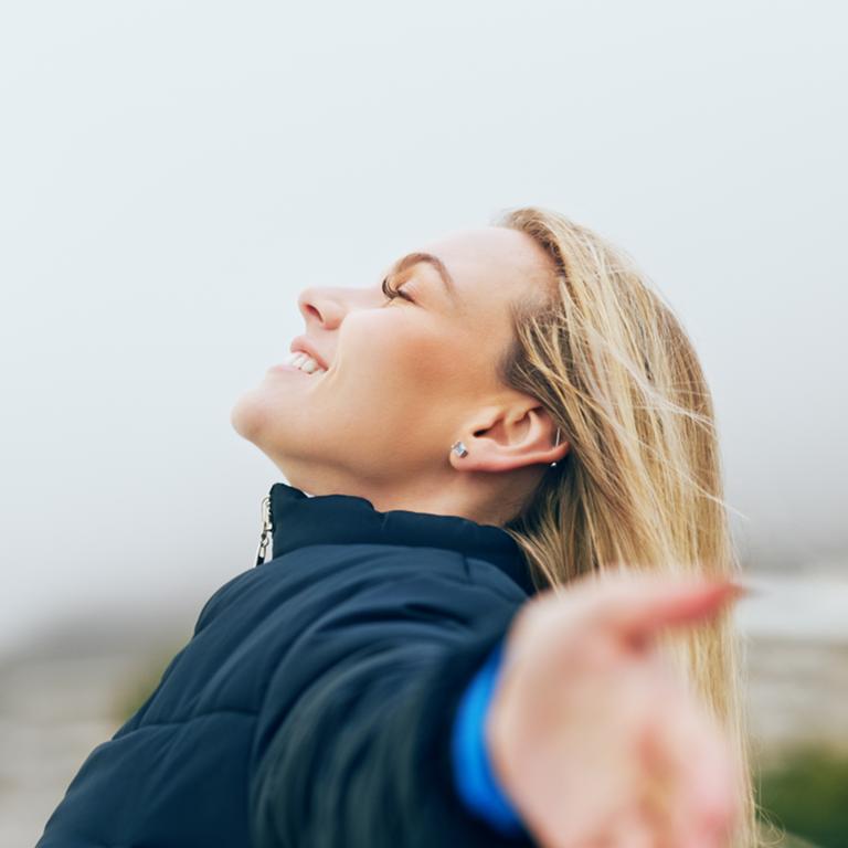 A woman standing outdoors with her arms outstretched, enjoying the fresh air, symbolising the importance of health and wellness in relation to oxygen insurance.