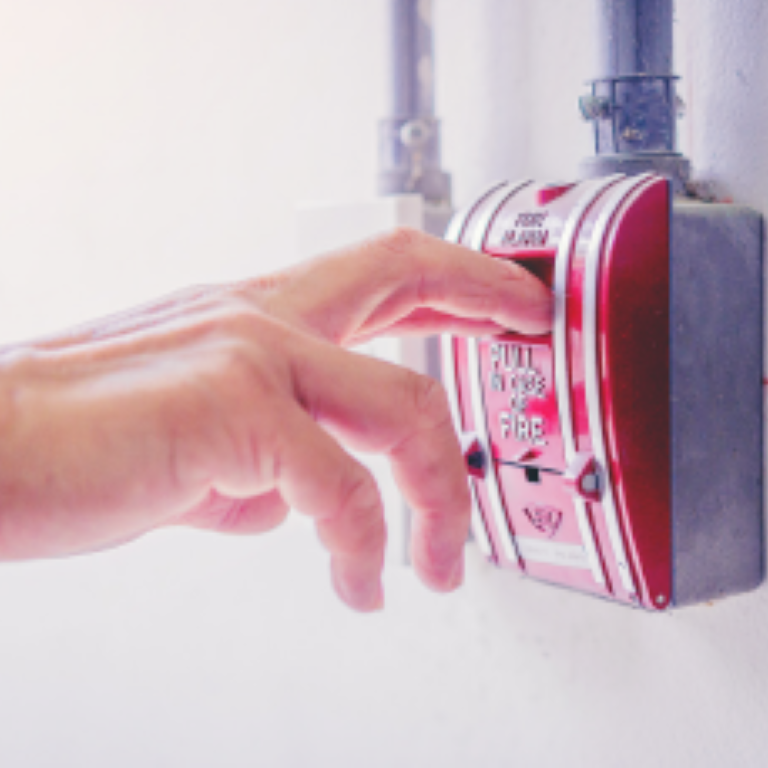 Fire safety for business leaders concept—hand pulling a wall-mounted fire alarm lever on a white wall.