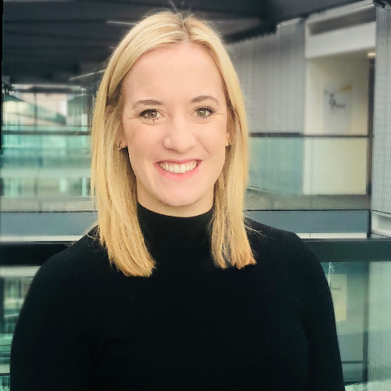 Robyn Beaman wearing a black high-neck top in a modern office setting, with shoulder-length blonde hair.