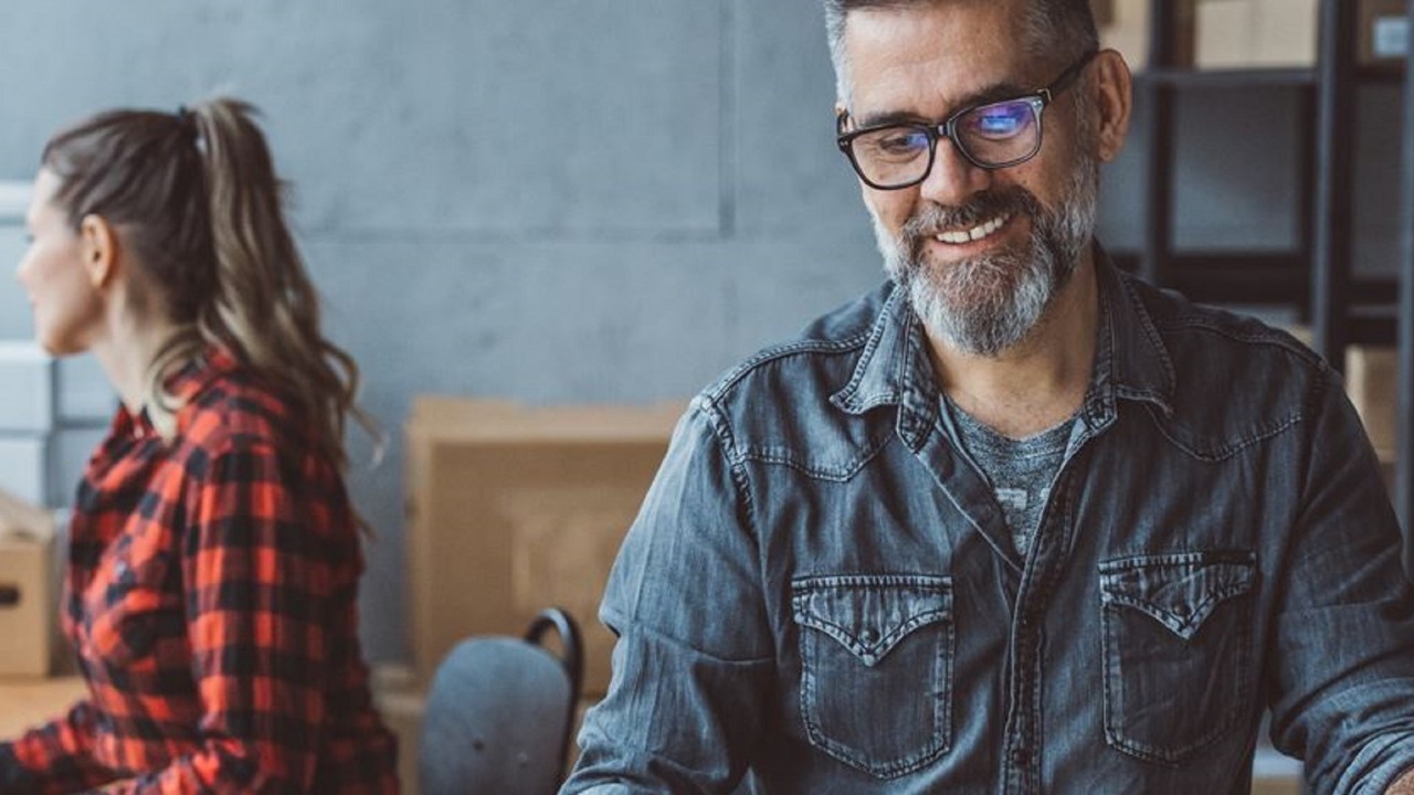 Ecommerce business owner smiling and working with co-worker, carboard boxes and shelving behind: ecommerce business insurance