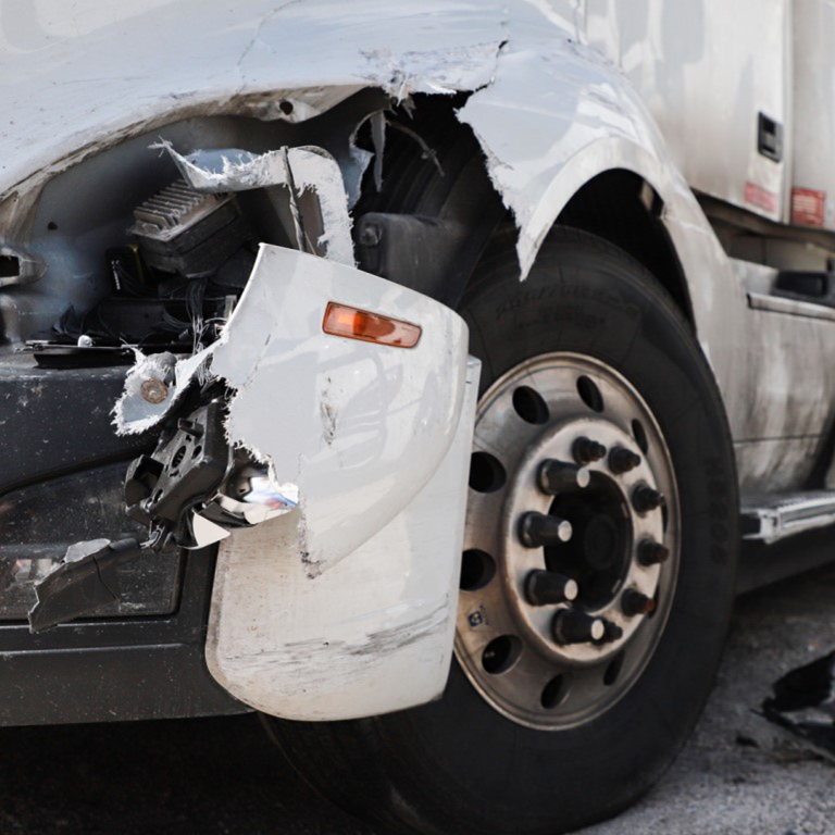 Close-up of a damaged white truck with a broken front bumper and debris on the ground beside the wheel.